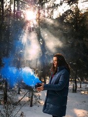 A young woman holds in her hands a vessel with developing blue smoke against the background of sunbeams in a winter forest.