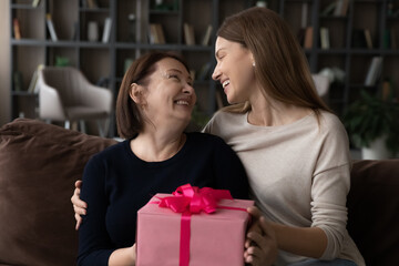 Loving young woman grown-up daughter congratulating mature mother, hugging, presenting pink gift...