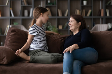Caring mature grandmother and little granddaughter talking, sitting on couch at home, happy senior woman with adorable girl child having fun, sharing secrets, telling news, enjoying leisure time