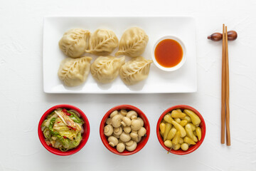 Boiled dumplings, manti or wontons with minced meat in a plate with sauce on a white table along with pickled mushrooms, hot peppers and cabbage salad. Homemade food. View from above