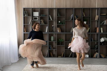 Happy woman with mature mother wearing pink princess skirts dancing having fun at home together,...