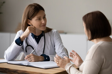 Female doctor therapist in white uniform with stethoscope listening to mature woman complaints at meeting, consulting, writing taking notes, filling patient history form, sitting at desk in hospital
