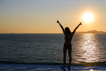 Silhouette of woman show hands happiness on the tropical sea beach near poolside in sunset time