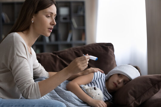 Caring worried mother checking sick little daughter high temperature, holding thermometer, little girl child with towel on forehead feeling unhealthy, having fever flu, lying on couch at home
