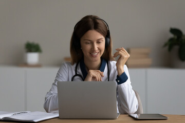 Smiling woman doctor in headphones consulting patient online, looking at laptop screen, young female practitioner therapist gp making video call or watching webinar, sitting at work desk in office