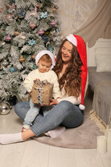 a young beautiful girl with long hair in a Santa Claus hat in a knitted white sweater jeans is sitting at the Christmas tree in the house in the arms of a little girl daughter in a Santa Claus hat