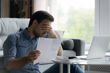 Depressed unhappy young man covering eyes holding paper document, feeling stressed of getting...