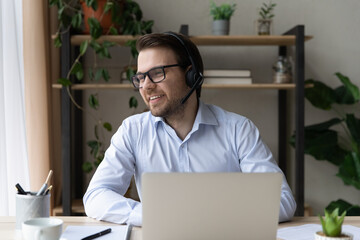 Happy dreamy young businessman in glasses wearing headphones with mic, looking in distance listening educational lecture or enjoying taking part in virtual event, distant communication concept.