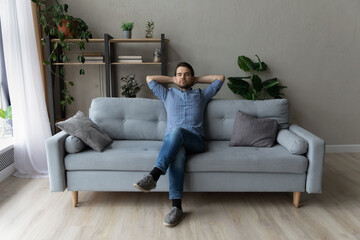Full length happy carefree millennial caucasian man relaxing on comfortable sofa, breathing fresh conditioned air, daydreaming napping enjoying peaceful weekend leisure time in modern living room.