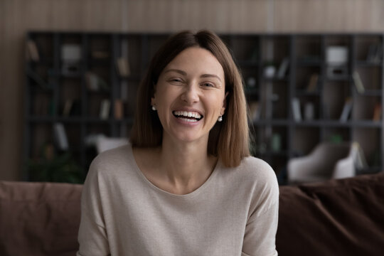 Head shot portrait of excited smiling woman looking at camera, chatting online by video call, talking and laughing, sitting on couch, happy young blogger shooting recording for social networks
