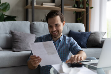 Happy millennial generation man managing holding paper bills, enjoying doing domestic paperwork, calculating monthly budget or expenses, making online payments in e-banking app, accounting concept.