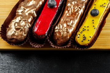 Multicolored glazed eclairs decorated with blueberries lie on a wooden cutting board.