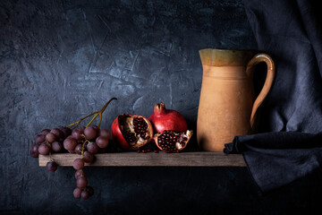 on the rustic wooden shelf, a bunch of grapes, pomegranates and an earthenware jug. Still life © luigi giordano
