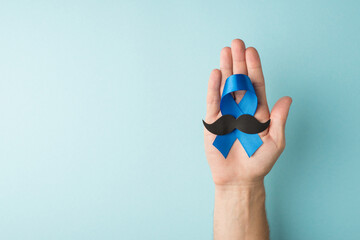 First person top view photo of male hand holding blue ribbon and mustache shape in palm symbol of prostate cancer awareness on isolated pastel blue background with empty space