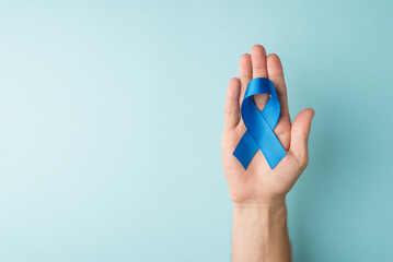 First person top view photo of male hand holding blue silk ribbon in palm symbol of prostate cancer awareness on isolated pastel blue background with blank space