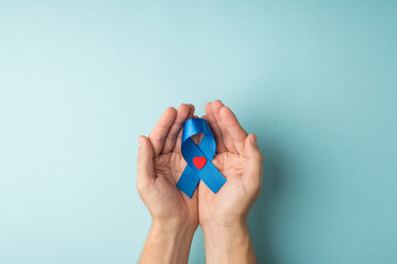 First person top view photo of man's hands holding blue ribbon with small red heart in palms symbol of prostate cancer awareness on isolated pastel blue background with copyspace