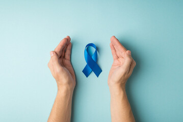 First person top view photo of blue ribbon symbol of prostate cancer awareness between male palms on isolated pastel blue background - obrazy, fototapety, plakaty
