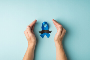 First person top view photo of blue ribbon and mustache shape symbol of prostate cancer awareness between male palms on isolated pastel blue background
