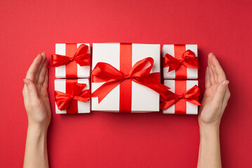 First person top view photo of white gift boxes with red ribbon bows between female hands on isolated red background