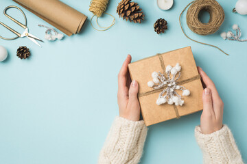 First person top view photo of woman's hands in knitted pullover holding giftbox decorated with snow twig and twine christmas tree balls handicraft tools isolated pastel blue background with copyspace