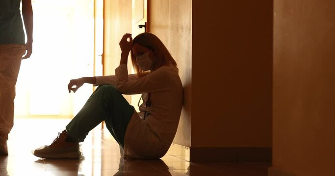 Sad doctor with protective medical mask sitting down in corridor of clinic 4k movie slow motion