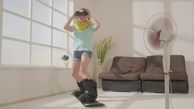 Woman in virtual reality glasses slide on snowboard in living room, simulated snowboarding using modern technology.