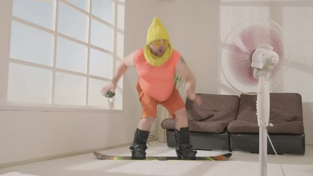 Funny fat man snowboarder fun snowboarding in living room of house.