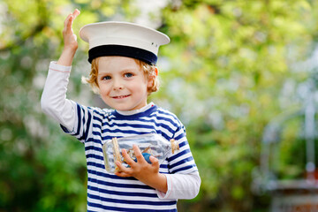 Happy little kid boy in sailor capitain hat and uniform playing with sailor boat ship. Smiling preschool child dreaming and having fun. Education, profession, dream concept - obrazy, fototapety, plakaty