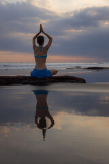 Sunset yoga. Caucasian woman sitting on the stone in Lotus pose. Padmasana. Hands in namaste mudra. Bali beach. View from back. Copy space. Yoga retreat. Water reflection. Healthy life. Zen life.