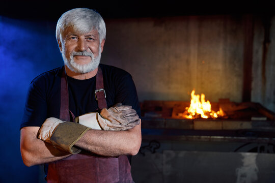 Caucasian blacksmith with grey hair and beard holding arms crossed near burning fire at forge. Portrait of aged industrial worker wearing protective apron and gloves. 