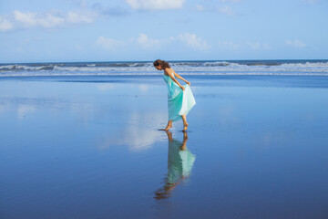 Young woman dancing barefoot on empty sandy beach. Full body portrait. Slim Caucasian woman wearing long dress. Water reflection. Summer vacation in Asia. Travel concept. Bali, Indonesia