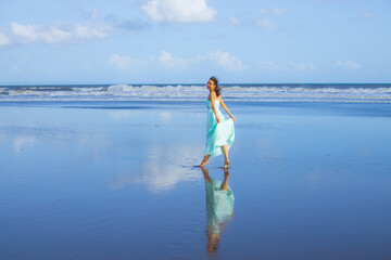 Happy smiling woman walking barefoot on empty beach. Full body portrait. Slim Caucasian woman wearing long dress. Water reflection. Blue sky. Vacation in Asia. Travel concept. Bali, Indonesia