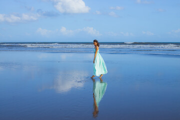 Happy smiling woman walking barefoot on sandy beach. Full body portrait. Slim Caucasian woman wearing long dress. Water reflection. Blue sky. Vacation in Asia. Travel concept. Bali, Indonesia