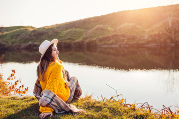 Woman relaxing by autumn lake at sunset. Stylish girl in hat sitting on bank enjoying fall landscape, breathing freely