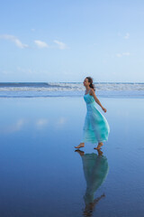Smiling woman walking barefoot on empty beach. Full body portrait. Slim Caucasian woman wearing long dress. Water reflection. Happiness and freedom. Summer vacation in Asia. Travel concept.