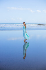 Happy smiling woman walking barefoot on empty beach. Full body portrait. Slim Caucasian woman wearing long dress. Water reflection. Blue sky. Vacation in Asia. Travel concept. Bali, Indonesia