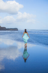 Young woman walking barefoot on empty beach. Full body portrait. Slim Caucasian woman wearing long dress. View from back. Water reflection. Blue sky. Travel concept. Bali, Indonesia