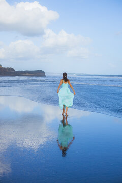 Young woman walking barefoot on empty beach. Full body portrait. Slim Caucasian woman wearing long dress. View from back. Water reflection. Blue sky. Travel concept. Bali, Indonesia