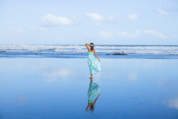 Young woman walking barefoot on empty beach. Full body portrait. Slim Caucasian woman wearing long dress. View from back. Water reflection. Blue sky. Travel concept. Bali, Indonesia