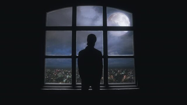 Male Silhouette Looking Full Moon Over City From Window, Zoom In. Silhouette of an isolated man looking to the full moon through a big window inside a house. Zoom in