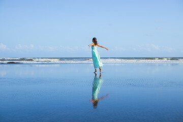 Young woman walking barefoot on empty beach. Full body portrait. Slim Caucasian woman wearing long dress. View from back. Water reflection. Freedom concept. Vacation in Asia. Bali