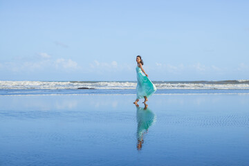 Young woman walking barefoot on empty beach. Full body portrait. Slim Caucasian woman wearing long dress. Enjoy time on the beach. Water reflection. Summer vacation in Asia. Travel concept. Bali