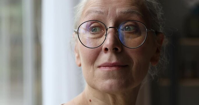 Close up face of sad upset elderly female pensioner in eyewear stand by window look aside sigh heavily lost in unpleasant thoughts. Unhealthy unwell older woman retiree grieve suffer alone has problem