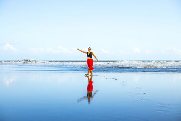 Happy woman walking barefoot on the beach. Full body portrait. Caucasian woman wearing red skirt. Enjoy time on the beach. Water reflection. Summer vacation in Asia. Travel concept. Bali