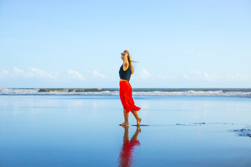 Young woman walking barefoot on the beach. Hands raised up. Caucasian woman wearing red skirt. Enjoy time on the beach. Water reflection. Summer vacation in Asia. Travel concept. Bali