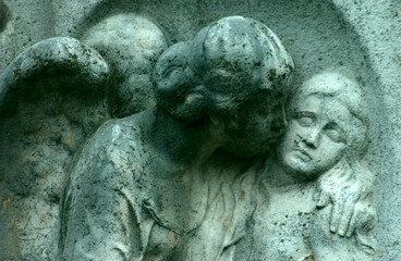 An old, weathered sandstone sculpture of an male angel who holds a young woman in his arms.