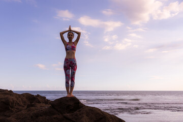 Slim woman standing on the rock, practicing yoga at the beach. Hands raised up in namaste mudra. Blue sky background. Yoga retreat. Concentration and balance. Horizontal layout. Copy space.