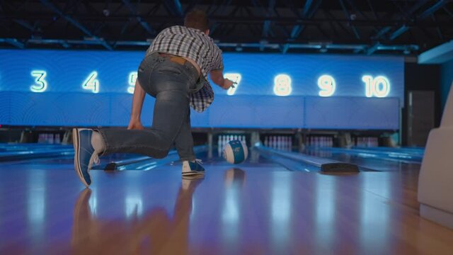 The camera follows one Caucasian man throwing a bowling ball on the playing track and jumping rejoicing in the downed pins. One man bowling in slow motion