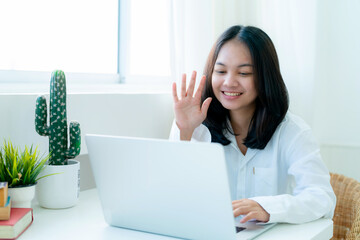 Portrait Cute Asian teen with long hair Wearing a white shirt, sitting online studying with a laptop computer on the table in the camera to say hello to friends through the camera.