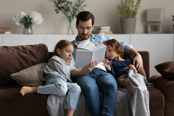 Caring father in glasses reading book to little daughter and son, hugging, sitting on cozy couch at home, family spending leisure time together, loving dad with kids engaged in educational activity